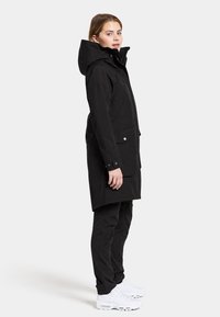 Cappotto lungo nero con cappuccio, tasche a doppio snap e dettagli ai polsini con snap, abbinato a pantaloni neri e sneakers bianche, su uno sfondo neutro.
