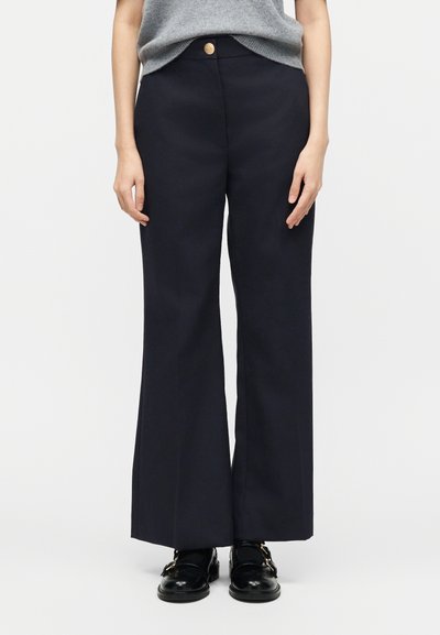 Femme portant un pantalon noir taille haute à jambes larges avec un bouton à l'avant, et des mocassins en cuir noir avec des boucles dorées.