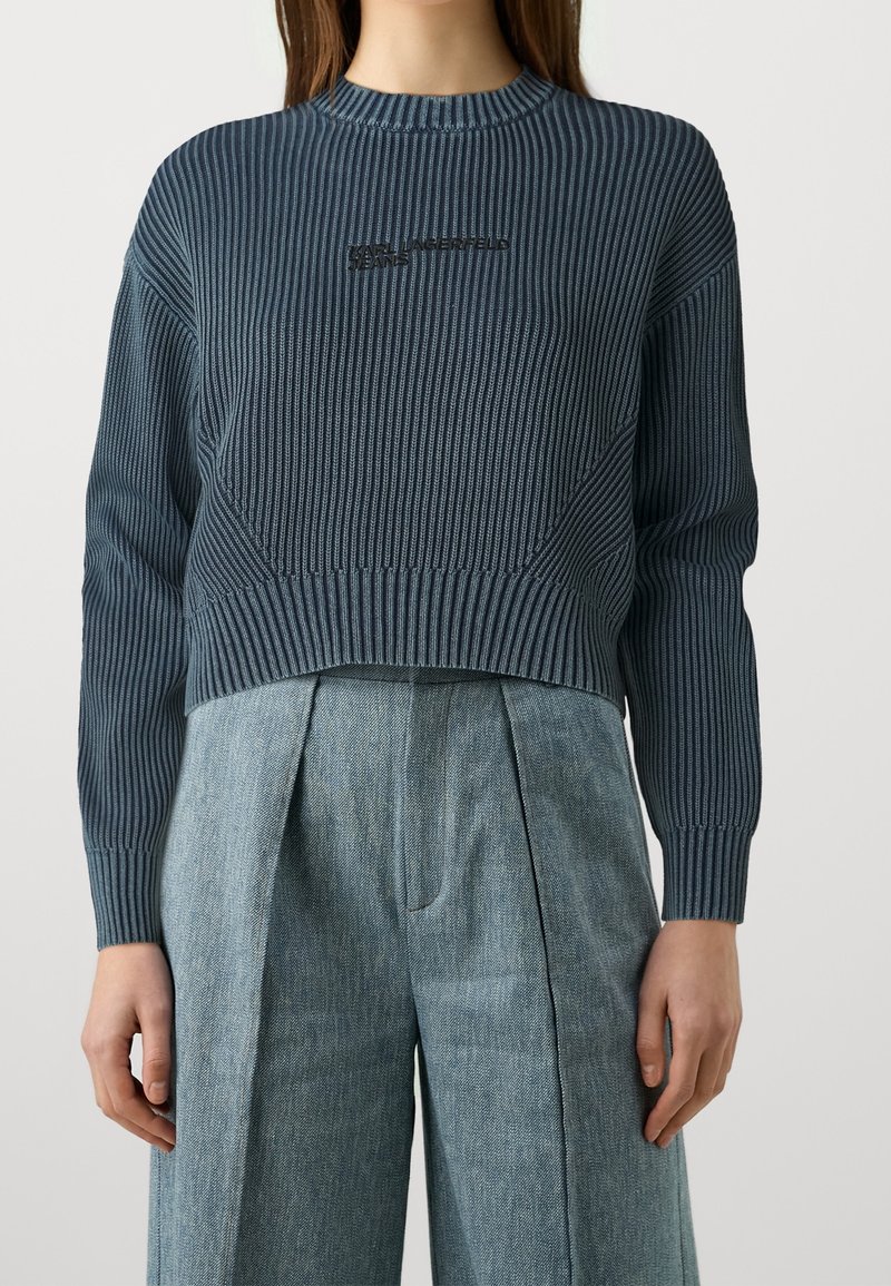 Femme portant un pull côtelé bleu foncé avec le texte « KARL LAGERFELD JEANS », associé à un pantalon taille haute à jambes larges bleu clair.