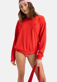 Red sweatshirt with long sleeves and a ribbed hem, featuring a subtle logo. Paired with striped bikini bottoms and a red bag.