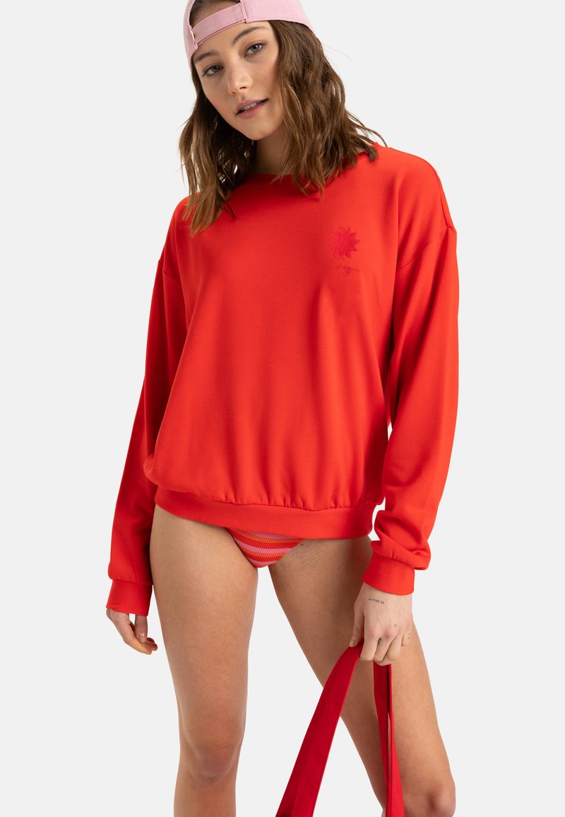 Red sweatshirt with long sleeves and a ribbed hem, featuring a subtle logo. Paired with striped bikini bottoms and a red bag.