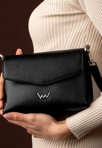 Černá kožená kabelka s texturovaným povrchem, stříbrné logo na přední straně a skládací klopa. Obsahuje odnímatelný ramenní popruh.