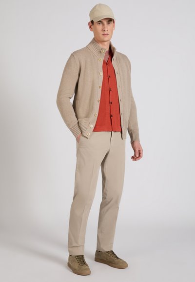Uomo che indossa un cardigan beige lavorato a maglia sopra una camicia color ruggine con bottoni, pantaloni beige, cappellino coordinato e sneakers marrone chiaro, in piedi con le mani nelle tasche.