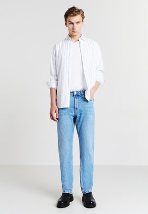 Giovane uomo in piedi con una mano in tasca, indossa una camicia bianca a righe, una maglietta bianca, jeans azzurri chiari e stivali neri su uno sfondo bianco.