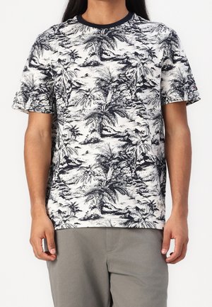 Mężczyzna w czarno-białej koszulce z krótkim rękawem z tropikalnym nadrukiem palm, w połączeniu z szarymi spodniami, stoi na białym tle.