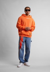 Oransje hettegenser med logo, blå jeans og hvite sneakers; rød bandana hengende fra jeanslommen; glatt stoff, avslappet passform.