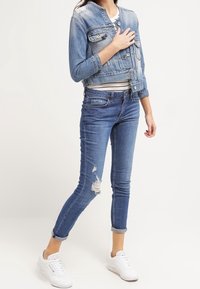 Kvinna som bär en kort denimjacka, trasiga skinny jeans med uppvikta benslut och vita sneakers, stående med ett ben något framåt.