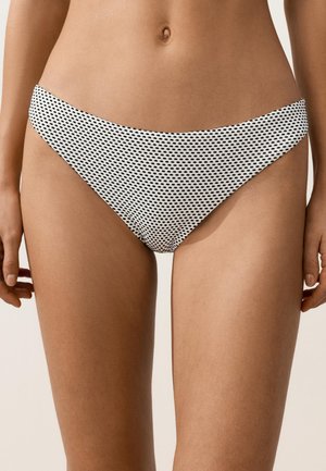 Donna che indossa slip bikini bianchi con piccoli motivi ovali neri, mostrata dalla vita alla metà della coscia su uno sfondo neutro.