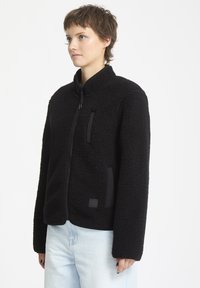 Giacca in fleece nera con colletto alto, chiusura con zip e tasche laterali. Tessuto testurizzato con una sensazione morbida e pelosa e dettagli sintetici.