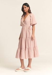 Ružová gingham midi šaty s hlbokým výstrihom do V, nariasenými rukávmi a vrstevnatou sukňou. Obsahujú opasok a sú vyrobené z mäkkej, ľahkej tkaniny.