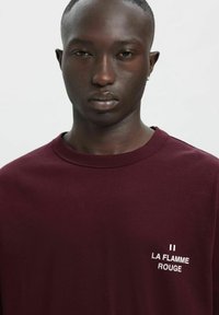 Bordo bombažna majica s posadkim ovratnikom, kratkimi rokavi in belim napisom "LA FLAMME ROUGE" na prsih. Gladek otip.