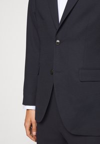 Dunkelblazer in Nachtblau mit strukturiertem Stoff, zwei Knöpfen und einer Brusttasche. Die Manschetten des Hemdes sind unter den Ärmeln sichtbar.