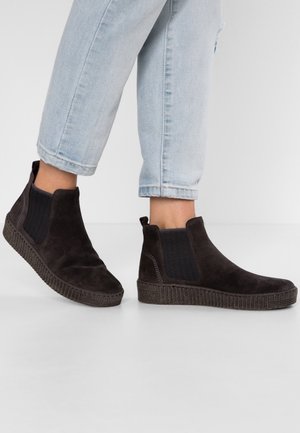 Person iført ankelhøje sorte ruskinds slip-on-støvler med elastiske sider og teksturerede såler, kombineret med lyseblå op rullede jeans.