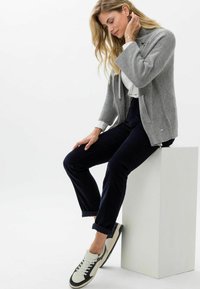 Grijze gebreide cardigan met een ritssluiting aan de voorkant, witte lange mouwen top, marineblauwe broek en witte sneakers. Model zittend op een wit voetstuk.