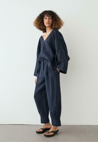 Žena s kudrnatými vlasy, oblečená v volném tmavě modrém tričku s proužky a výstřihem do V a odpovídajících zúžených kalhotách, stojící naboso na šedé podlaze u bílé zdi.