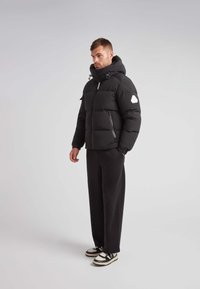 Giubbotto a sacco nero con colletto alto, chiusura zip frontale e polsini a costine. Presenta un cappuccio removibile e una patch con logo bianco sulla manica.