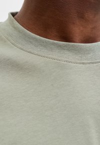 Nahaufnahme des Halses einer dunkelhäutigen Person, die ein hellgraues Baumwoll-T-Shirt mit Rundhalsausschnitt trägt.