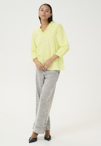 Blusa verde lime con scollatura a V e colletto a volant, con maniche lunghe e texture a zigzag. Abbinata a jeans a gamba larga grigio chiaro.