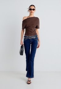 Καφέ τοπ με εκτεθειμένους ώμους, μπλε τζιν καμπάνα, μαύρη ζώνη με καρφιά, μαύρα γυαλιά ηλίου και μικρή μαύρη τσάντα με πουά.