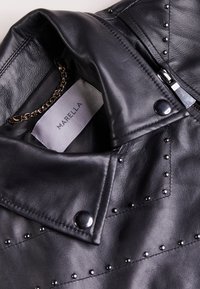 Zwarte leren jas met ritssluiting, studs op de kraag en het lijf, en een kettingaccent aan de binnenkant, met een label waarop "MARELLA" staat.