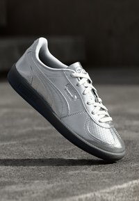 Sneaker synthétique argenté avec des panneaux texturés, bout arrondi, lacets blancs et semelle en caoutchouc noir. Présente le logo Puma sur le côté.
