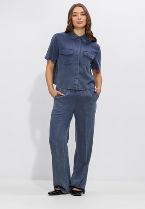 Frau steht mit Händen in den Taschen, trägt ein locker sitzendes, dunkelblaues kurzärmeliges Hemd mit Knöpfen und passend weit geschnittene Hose sowie schwarze Ballerinas.