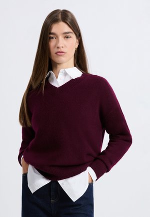 Pull en maille bordeaux avec un col en V, manches longues, poignets et ourlet côtelés. Porté sur une chemise blanche à col, associé à un jean denim foncé.