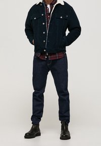 Donkerblauwe corduroy jas met een witte fleece kraag, gecombineerd met een rood geruit overhemd en marineblauwe spijkerbroek. Zwarte enkellaarsjes maken de outfit compleet.