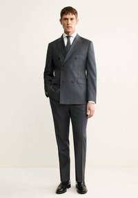 Completo grigio doppiopetto con finitura texturizzata, pantaloni slim-fit e una camicia bianca con cravatta nera; scarpe lucide completano il look.