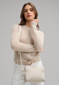 Beige crossbody taske med et tekstureret mønster og guld hardware. Båret med en cremefarvet striktrøje og lyse bukser. Justerbar rem.