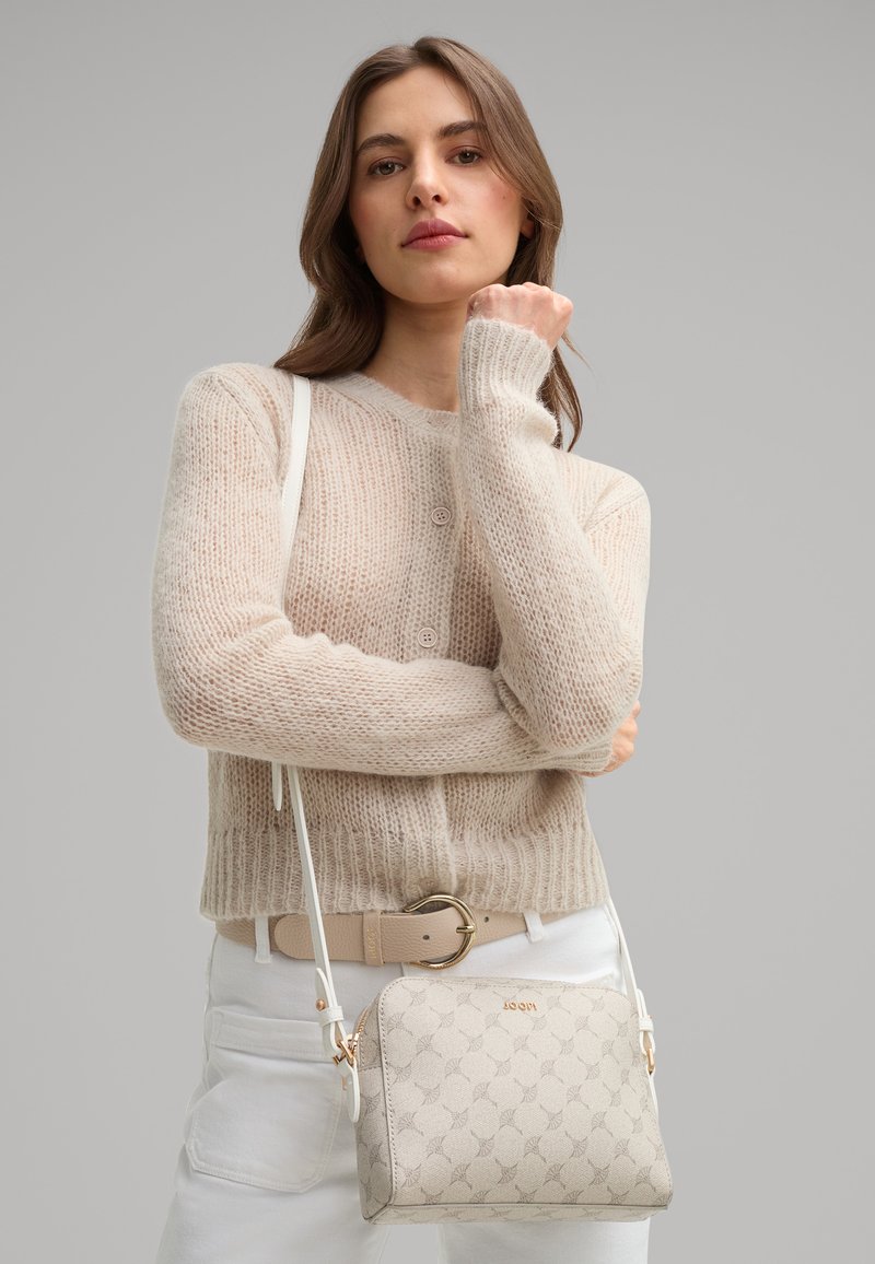 Beige crossbody taske med et tekstureret mønster og guld hardware. Båret med en cremefarvet striktrøje og lyse bukser. Justerbar rem.