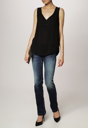 Kobieta ubrana w czarną bezrękawnikową bluzkę, wyblakłe niebieskie dżinsy, czarne szpilki, trzymająca czarną kopertówkę na neutralnym tle.