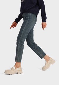 Mocassini beige con plataforma e accenti bianchi, dotati di suola spessa e texture in pelle liscia, indossati con jeans fitted blu scuro.