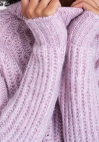 Ljusrosa stickad tröja med ett strukturerat mönster. Har ribbade ärmmuddar och en rullad krage, tillverkad av mjukt, fluffigt garn.