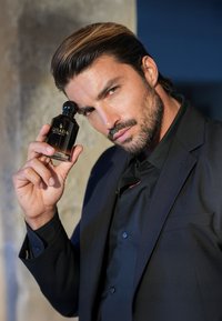 Czarna szklana butelka perfum z zaokrągloną zakrętką, opatrzona złotym napisem „GISADA”. Osoba trzyma ją blisko twarzy, ubrana w czarną koszulę.