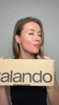 Woman with long hair holds a Zalando cardboard box, showing a slight smile, against a plain gray background.