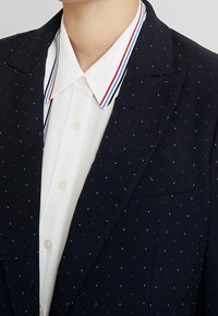 Blazer marine avec de petits points rouges et bleu clair, accompagné d'une chemise blanche avec un col rayé multicolore. Tissu texturé, coupe ajustée.