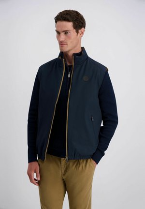 Mann trägt marineblaue ärmellose Weste mit Reißverschluss über dunklem Langarmpullover und hellbraunen Hosen, steht mit teilweise in den Taschen steckenden Händen.