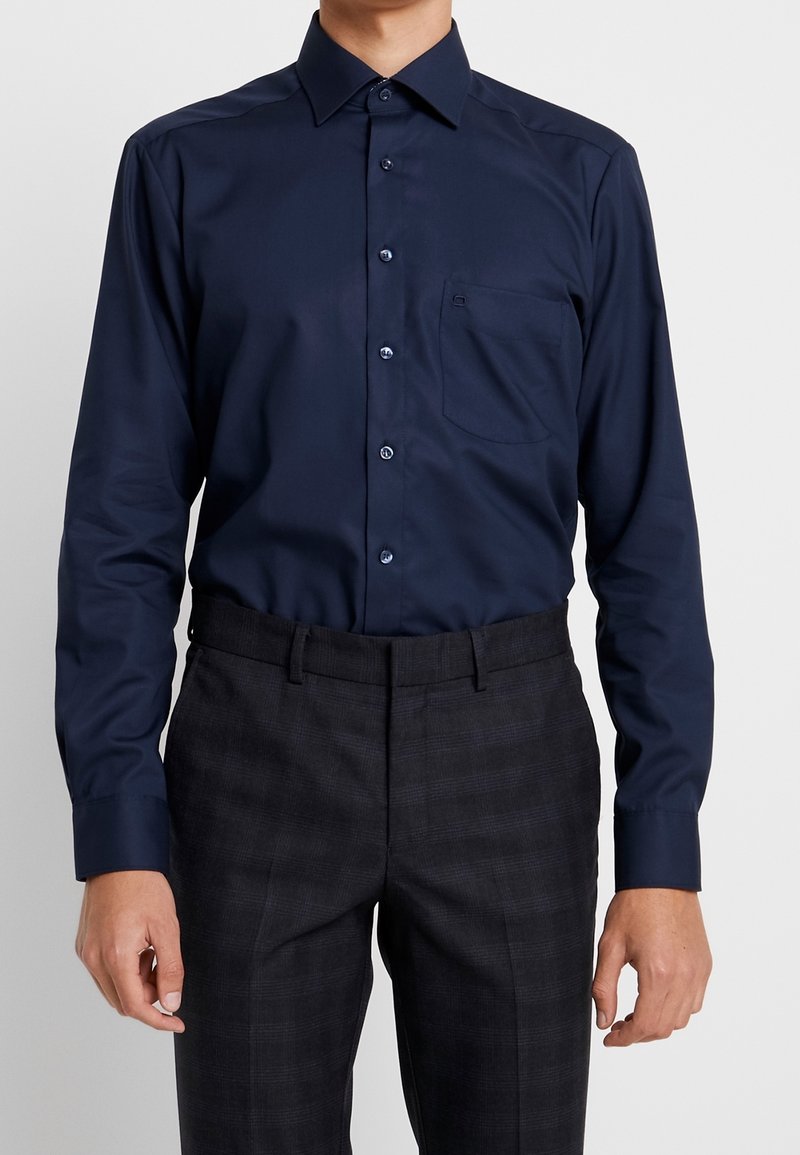 Homme portant une chemise bleu foncé à manches longues avec une poche poitrine, rentrée dans un pantalon noir sur mesure avec un motif subtil.