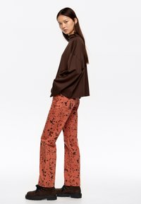 Ruda didelė megztinis ilgomis rankovėmis, derinamas su koralinėmis kelnėmis, turinčiomis juodų purslų raštą, ir rudais auliniais batais.