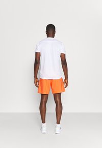 Camisa blanca de manga corta y pantalones cortos naranja brillante, hechos de una tela ligera. Zapatillas deportivas blancas con acentos sutiles, vistas desde atrás.