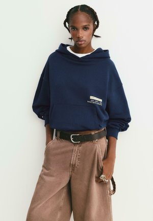 Jeune femme avec des cheveux tressés portant un sweat à capuche navy avec du texte et un pantalon large marron, se tenant avec une main dans la poche sur un fond uni.