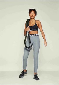 Moteris, vilkinti juodą sportinį braletą ir pilkas Nike leggingas, laiko suktą kovos virvę ant peties, stovėdama prie vientiso fono.