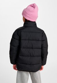 Barn vendt væk iført en sort polstret jakke, lyserød strikket hue og lysegrå sweatpants mod en plain hvid baggrund.