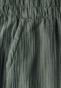 Tessuto verde testurizzato con righe verticali, dotato di una vita elasticizzata e di un piccolo tag metallico del marchio. Materiale morbido e leggero.