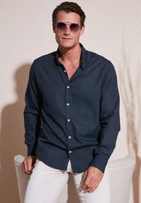 Camicia button-up blu scuro con maniche lunghe, realizzata in tessuto testurizzato, bottoni bianchi, colletto parzialmente sbottonato, abbinata a pantaloni bianchi.