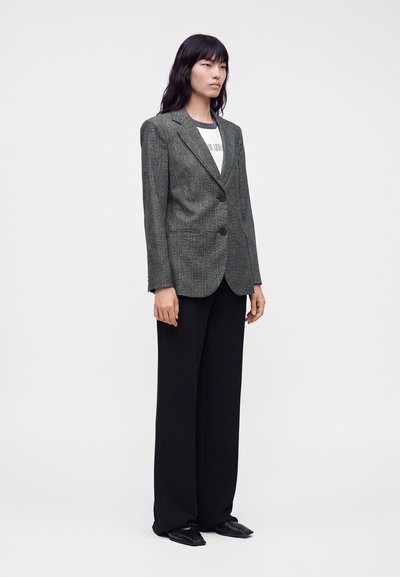 Femme aux longs cheveux noirs portant un blazer à carreaux gris sur une chemise blanche, un pantalon noir à jambes larges et des chaussures noires, debout devant un fond blanc.