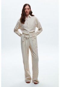 Outfit color crema con righe verticali, composto da una camicia con bottoni e pantaloni con coulisse. Entrambi presentano un tessuto leggero e strutturato.
