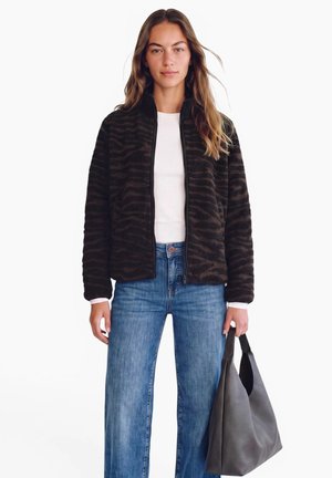 Junge Frau trägt eine dunkel gemusterte Jacke mit Tiermotiv, weißes Hemd und blaue Jeans, hält eine große graue Handtasche in der rechten Hand.