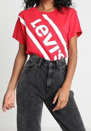 Kobieta w czerwonym t-shirtcie Levi's włożonym do czarnych wysokich, spranych dżinsów, stoi z jedną ręką blisko biodra, a drugą zrelaksowaną.